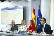 La imagen muestra una rueda de prensa o una presentación oficial del Gobierno de España. En la mesa hay cuatro personas vestidas formalmente, con documentos y micrófonos frente a ellos.  Detrás de los ponentes, se pueden ver dos banderas: la de España y la de la Unión Europea. En la pantalla de presentación se muestran gráficos con datos económicos, lo que sugiere que están hablando sobre la situación económica del país o presentando previsiones macroeconómicas. 