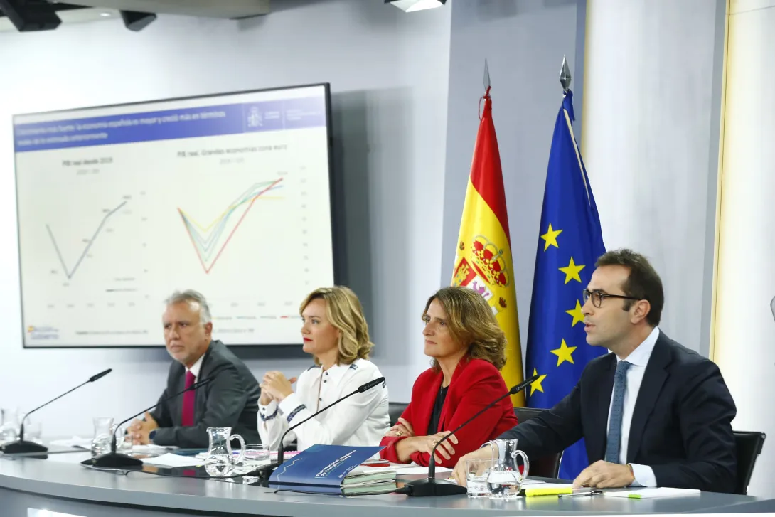 La imagen muestra una rueda de prensa o una presentación oficial del Gobierno de España. En la mesa hay cuatro personas vestidas formalmente, con documentos y micrófonos frente a ellos. Detrás de los ponentes, se pueden ver dos banderas: la de España y la de la Unión Europea. En la pantalla de presentación se muestran gráficos con datos económicos, lo que sugiere que están hablando sobre la situación económica del país o presentando previsiones macroeconómicas.
