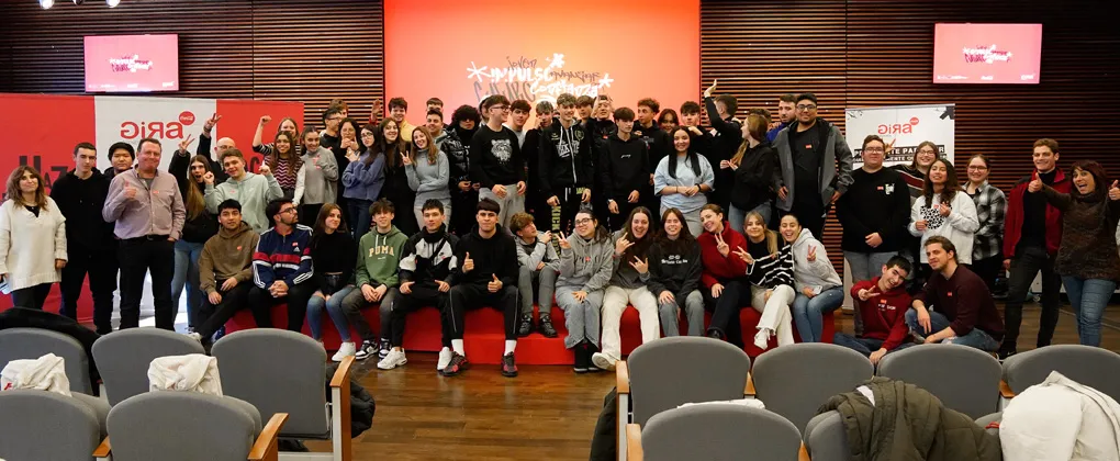 Un grupo numeroso de jóvenes posa en un auditorio tras una sesión del programa GIRA Jóvenes de Coca-Cola. Están de pie y sentados en un escenario con un fondo rojo que muestra el logo del evento. Muchos sonríen, levantan el pulgar o hacen gestos de entusiasmo. A los lados, algunos mentores y organizadores del programa los acompañan. En el fondo, se ven pantallas y carteles con el nombre del evento