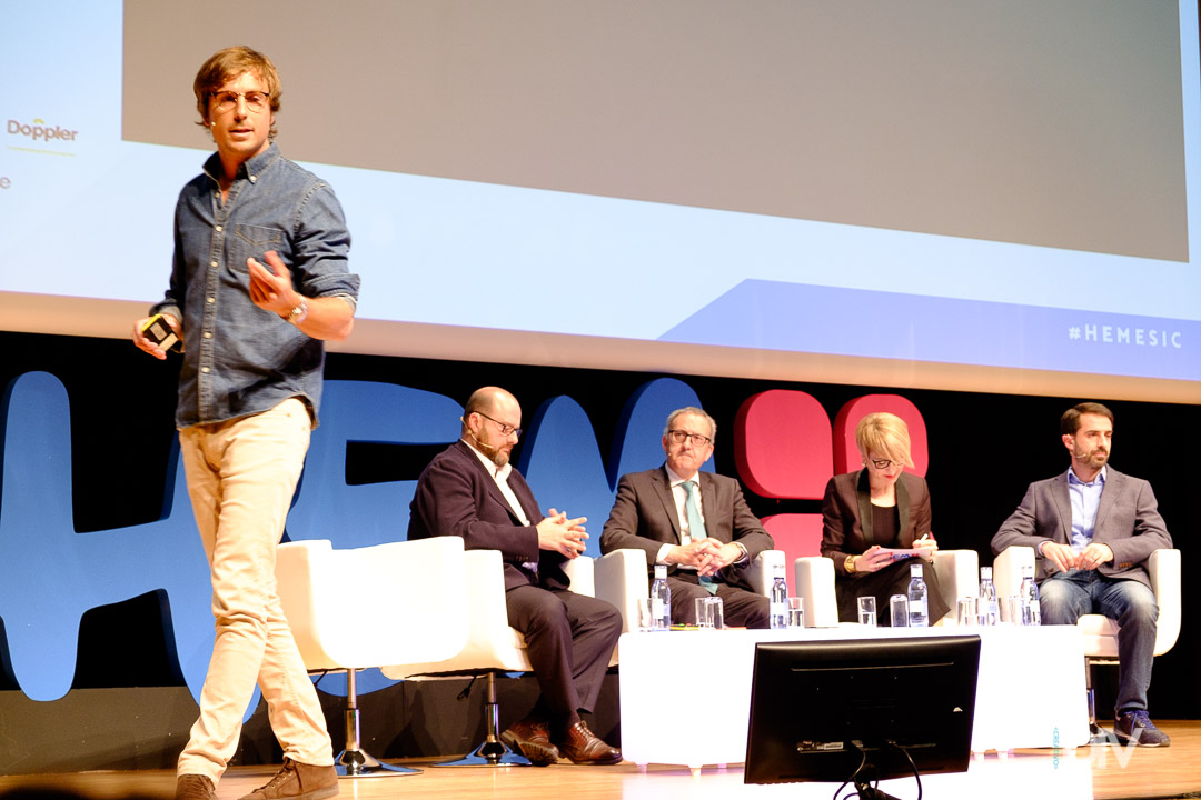 Un hombre joven con gafas y camisa azul camina por el escenario mientras habla en un evento. Detrás de él, cuatro panelistas con vestimenta formal están sentados en sillas blancas, con un gran cartel de fondo. Dos de ellos revisan documentos mientras los otros observan con atención.